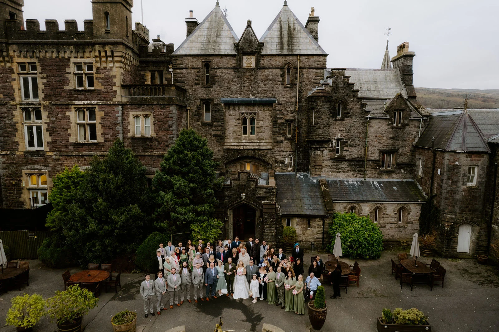 An aerial view of the wedding party gathered in the courtyard in front of Craig y Nos Castle, a grey stone castle with turrets and gables