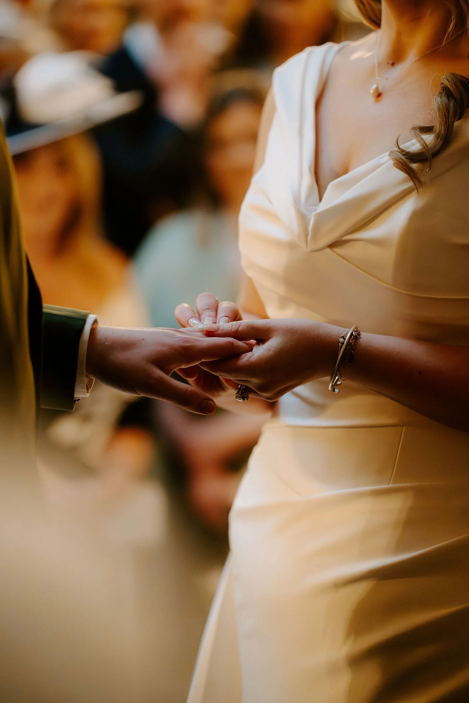 Close view of a groom sliding a ring onto a bride's finger during the ceremony, with seated guests softly blurred in the background.