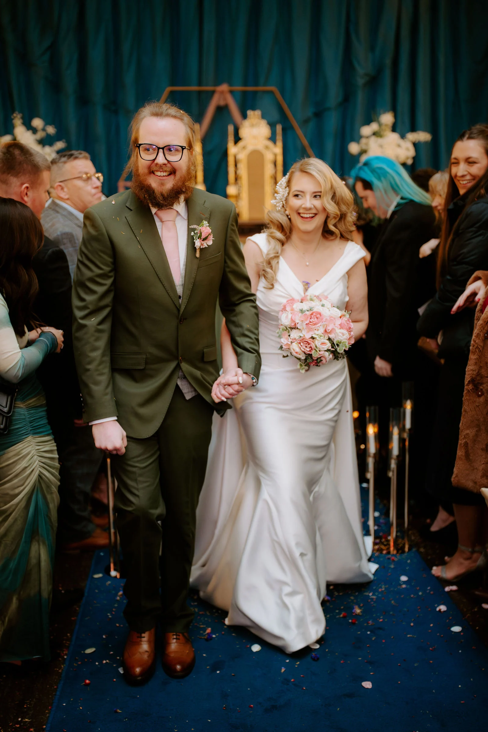 Tom in a green suit and Sophie in a satin gown holding a peach bouquet walk back through cheering guests on a blue carpet inside the castle