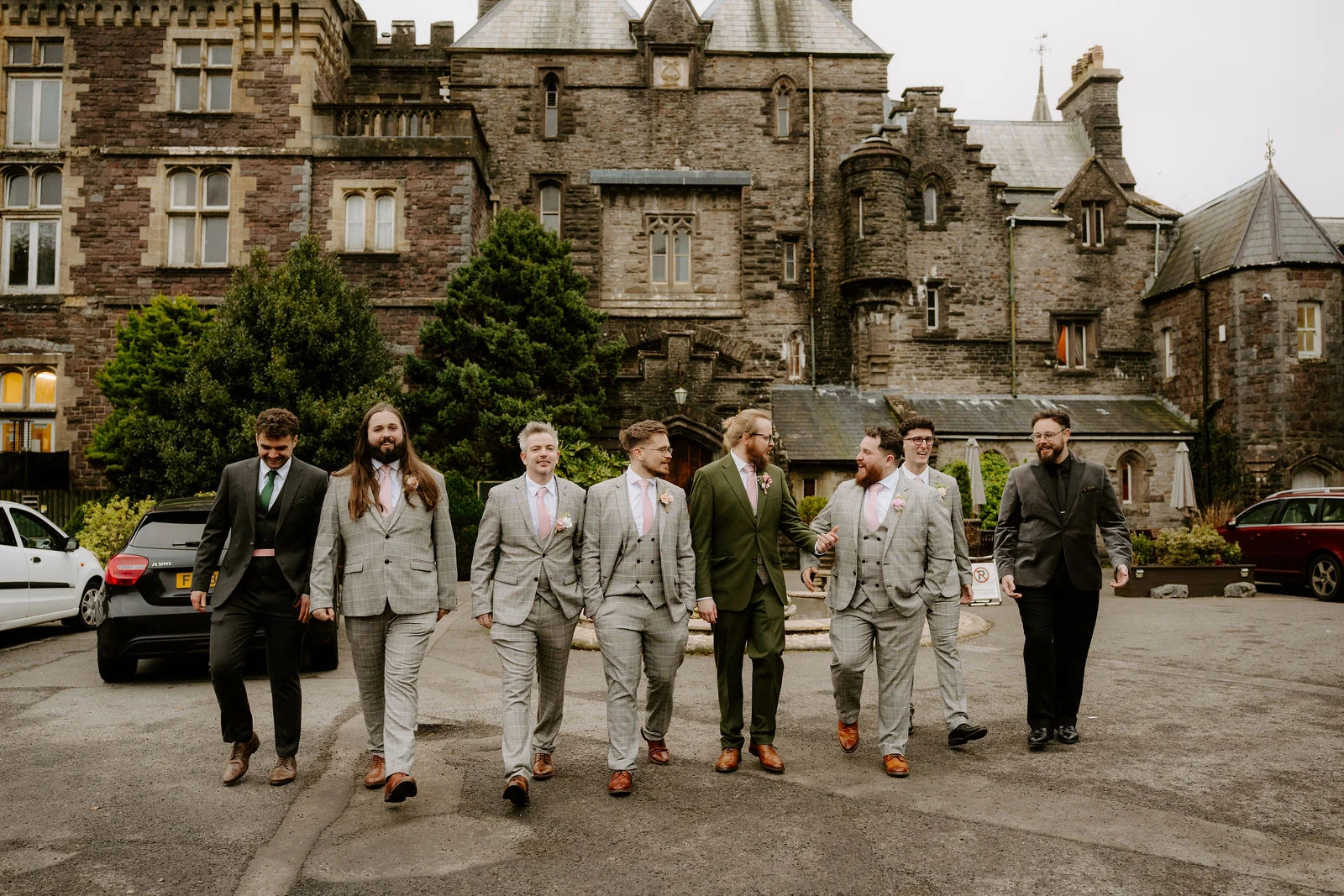 Eight groomsmen in grey and dark green suits walk side by side across a gravel courtyard, laughing, with the stone castle and its turrets rising behind them.