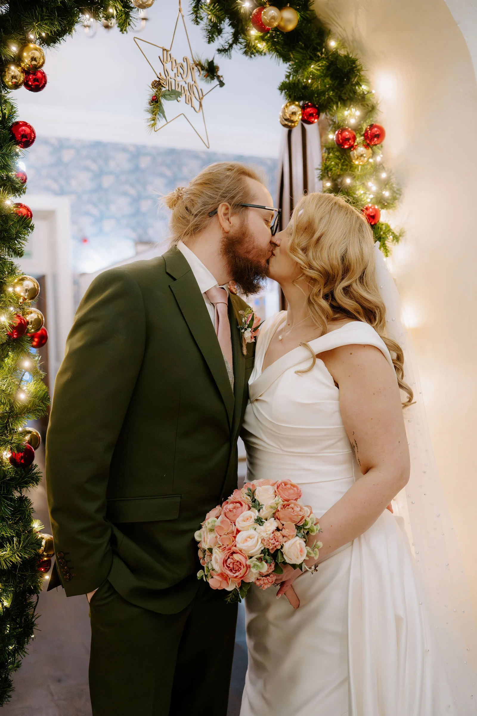 A groom in an olive green suit and a bride in an off the shoulder white dress share a kiss in front of a stone archway wrapped in a Christmas garland with lights and red and gold baubles.