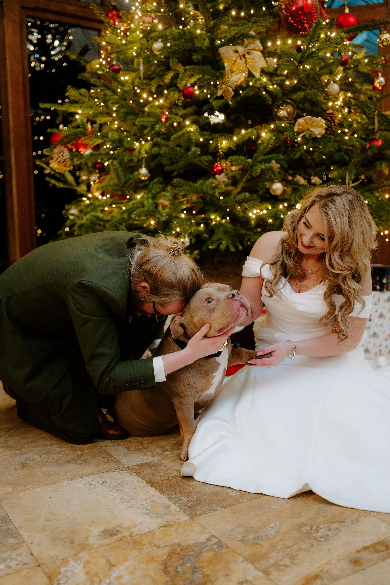 A groom in an olive green suit crouches to nuzzle a brown dog while a bride in a white dress sits on the stone floor beside them, smiling, in front of a lit Christmas tree.