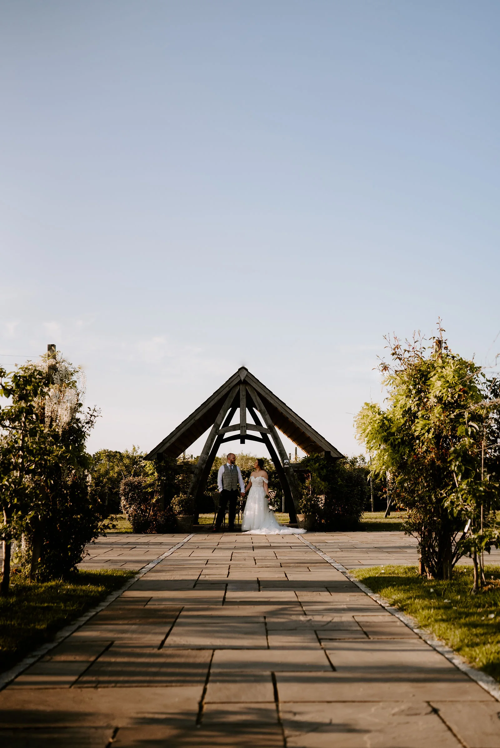 A distant view of a couple standing at the end of a paved path beneath a timber-framed pavilion, with trimmed hedges either side and a clear sky above