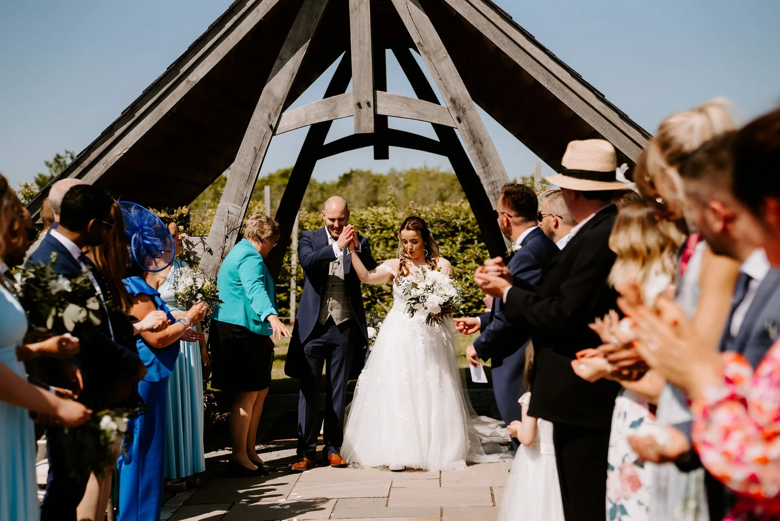 A bride in a lace gown with a bouquet and a groom in a waistcoat walk out beneath an open timber-framed pavilion as guests on both sides clap