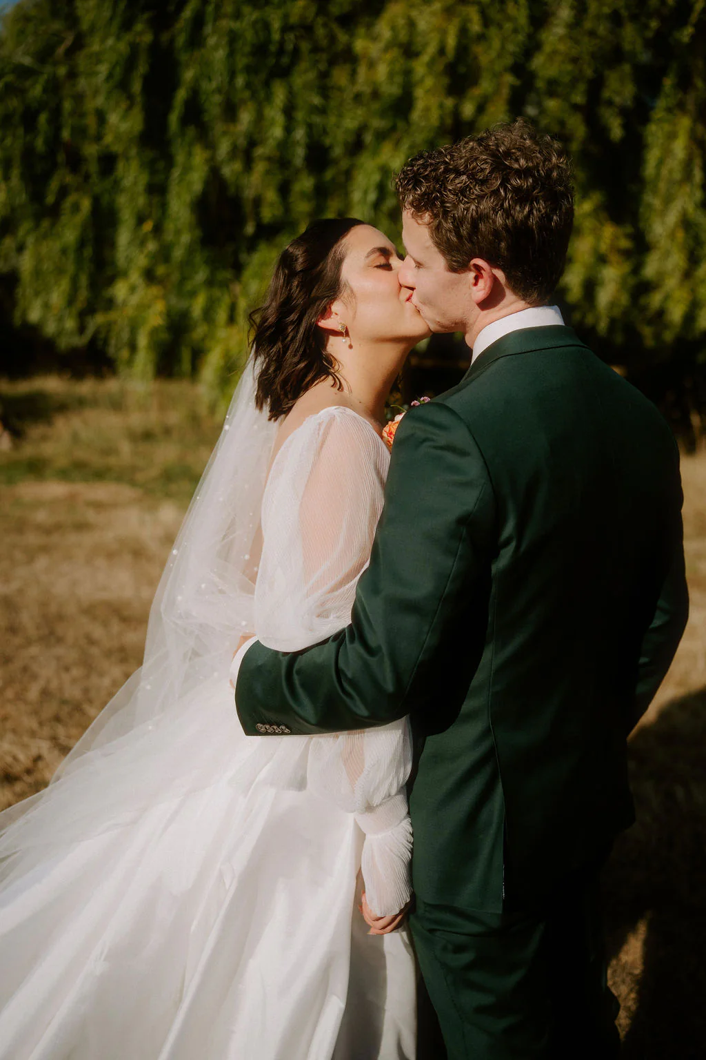 Taylor and Brodie kiss on the lawn, Brodie in a long sleeved gown and veil, with weeping willows behind them