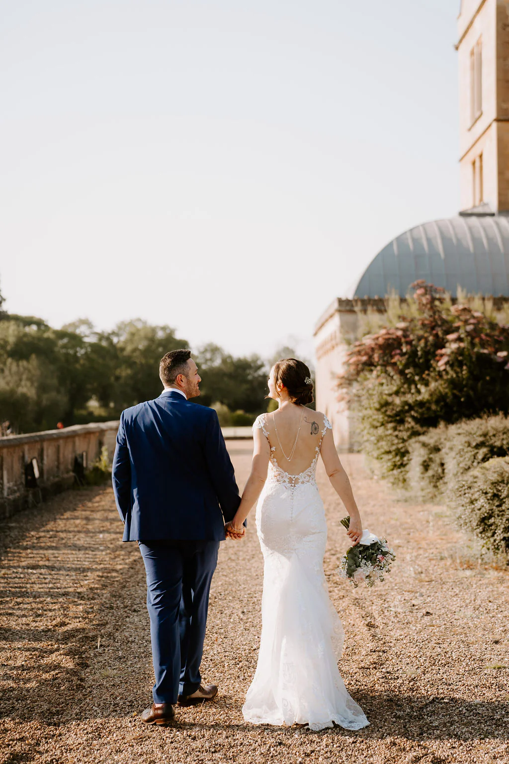 Kate and Josh walk hand in hand away from the camera along a gravel terrace beside a stone balustrade at Walton Hall in the evening sun