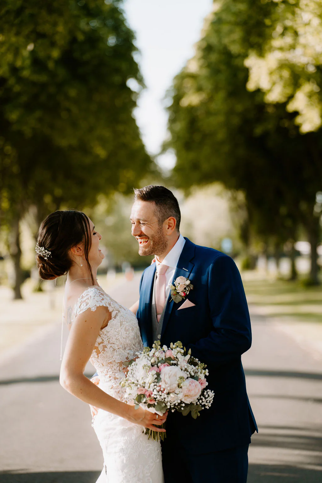 Kate and Josh smile at each other beneath an avenue of tall trees, Kate holding a pastel bouquet