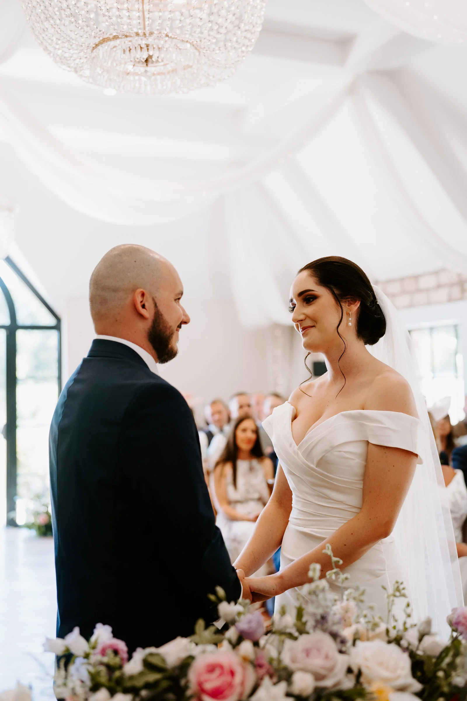 A bride in an off-the-shoulder gown and veil and a groom hold hands and look at each other beneath a crystal chandelier and white drapes, with a bouquet of pink and cream roses in the foreground and guests seated behind