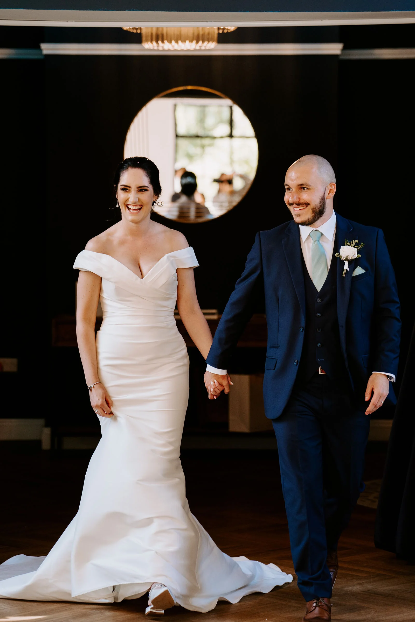 A laughing bride in a fitted gown and a groom in a navy three-piece suit walk hand in hand through a dark blue room with a round mirror on the wall