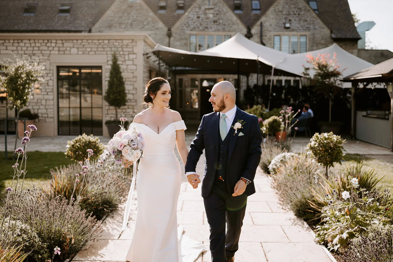 A bride holding a pink and cream bouquet and a groom in a navy suit walk hand in hand along a stone path between lavender borders, with a stone building and a stretch tent behind them