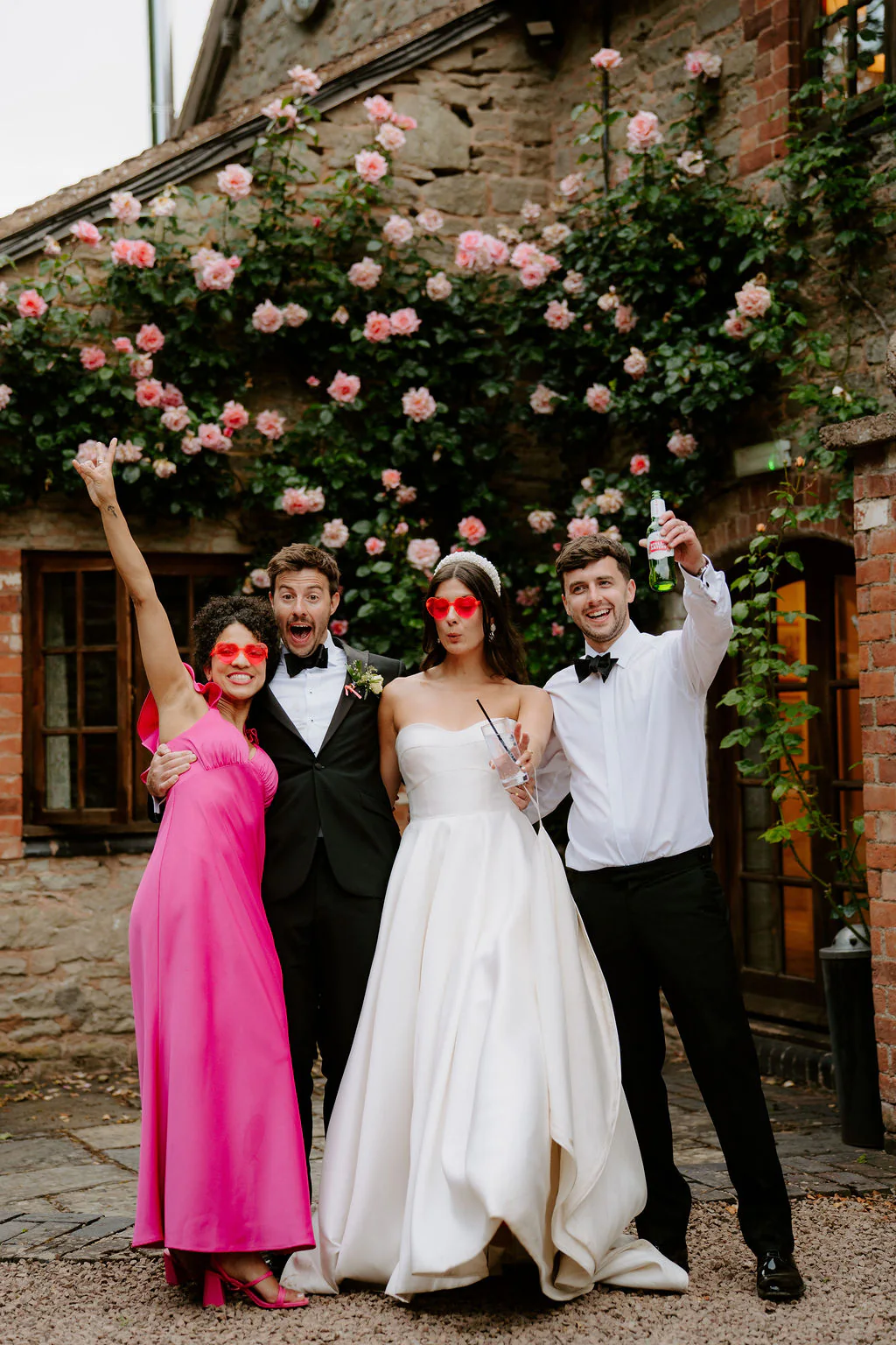 A groom, a bride, a bridesmaid in hot pink and a man in a white shirt cheer with their arms raised in front of a brick wall covered in pink roses
