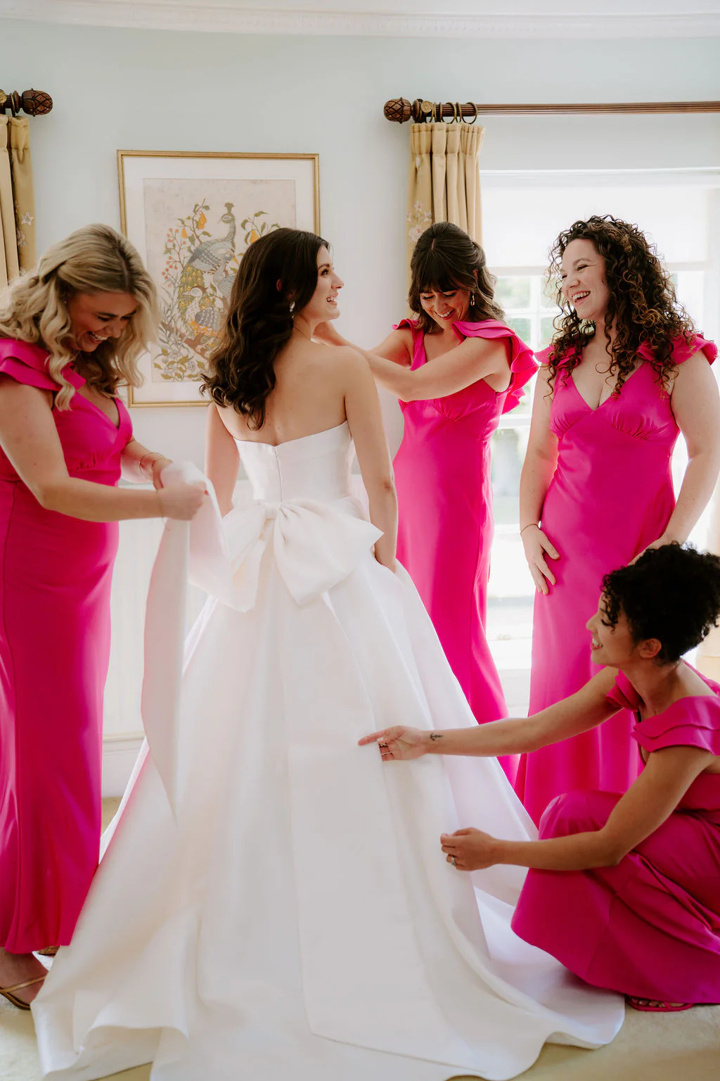 Bridesmaids in bright pink dresses help Jade into her ballgown beside a tall window at Birtsmorton Court