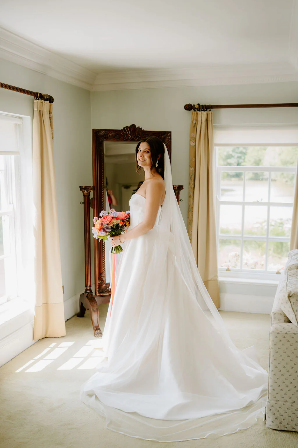 Jade in a veil and ballgown holds a bouquet in front of a tall mirror in a bright room at Birtsmorton Court