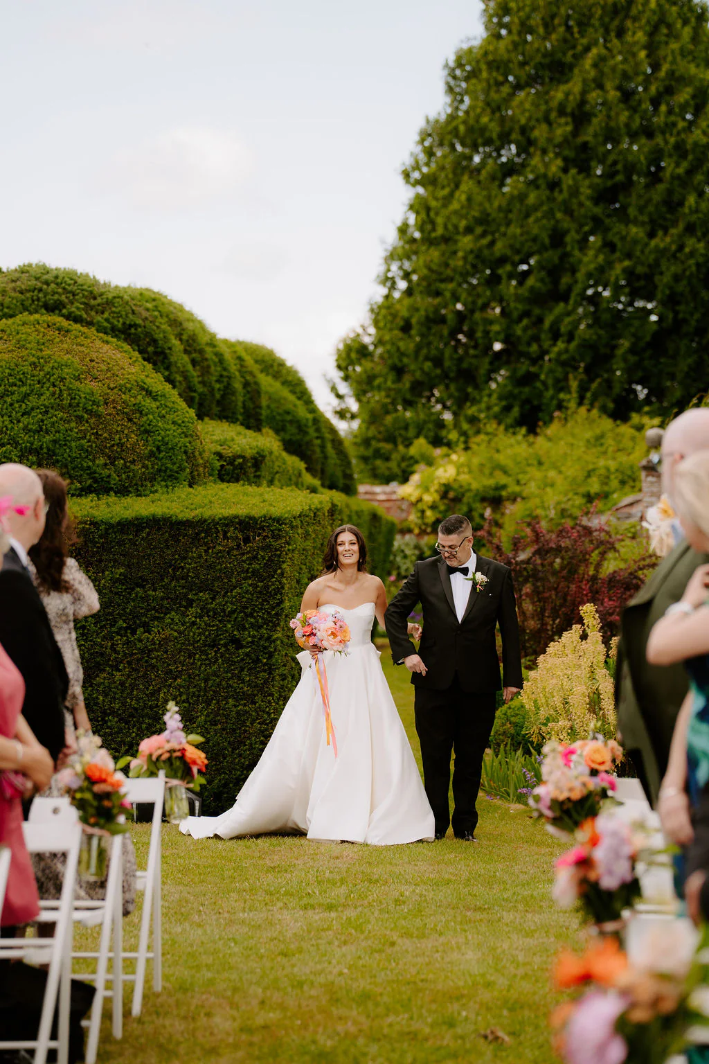 A bride and groom walk along a lawn aisle between rows of white chairs, with a tall clipped hedge ahead of them and guests either side