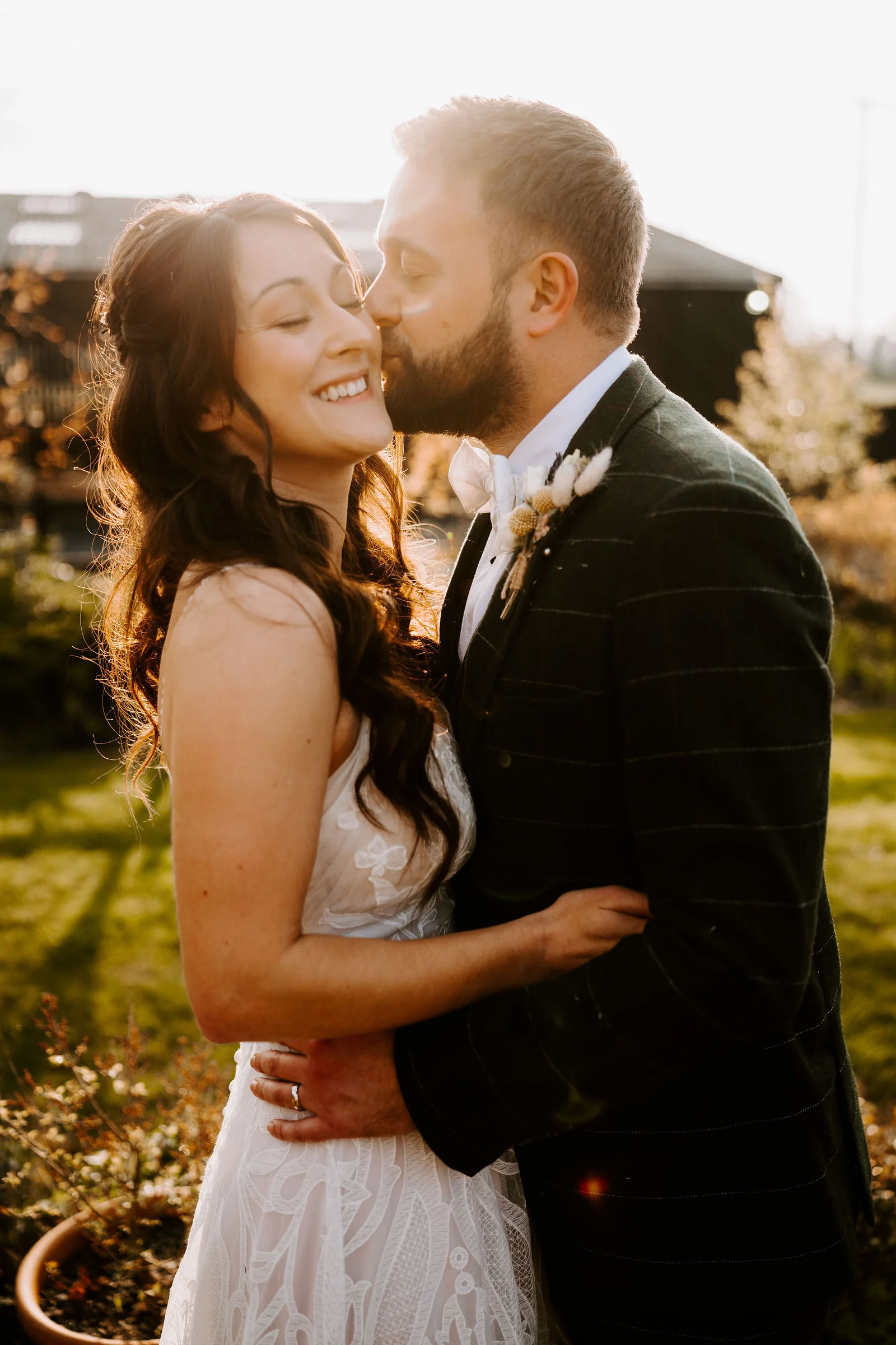 A groom in a dark check suit kisses the cheek of his laughing bride, backlit by low golden sun, with a dark timber barn behind them