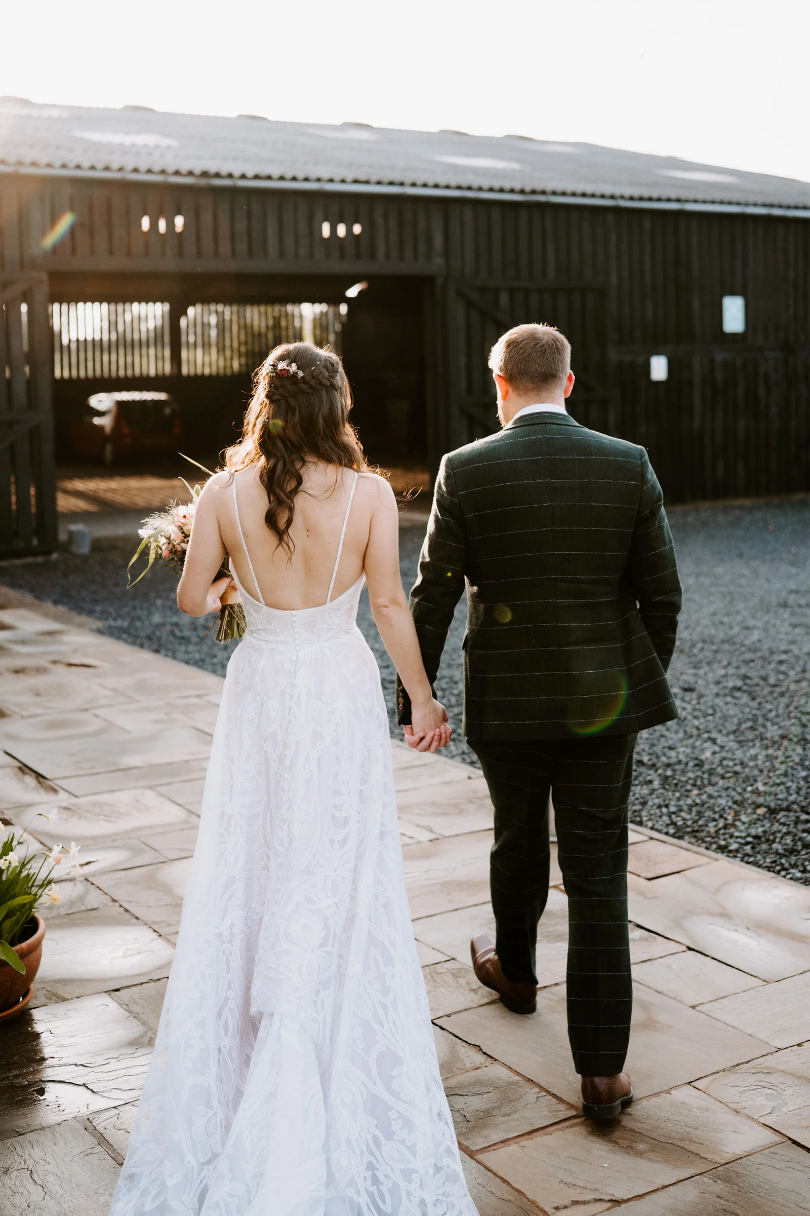 Seen from behind, a bride in a lace gown with an open back and a groom in a green check suit walk hand in hand across a paved yard towards a dark timber barn with open doors