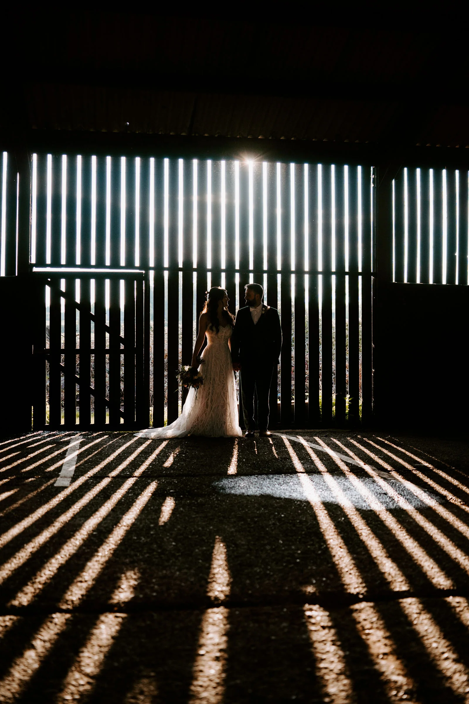 The silhouettes of a couple standing inside a dark barn as bright light through the vertical slats of its doors throws stripes across the floor