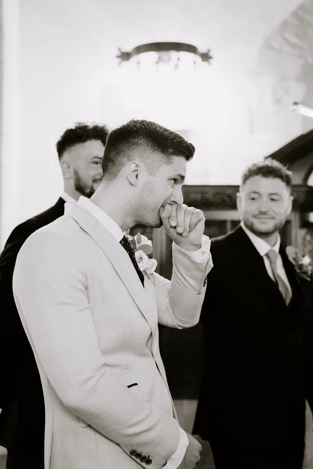 In black and white, a groom in a light suit wipes a tear from his eye with his hand while a groomsman smiles beside him