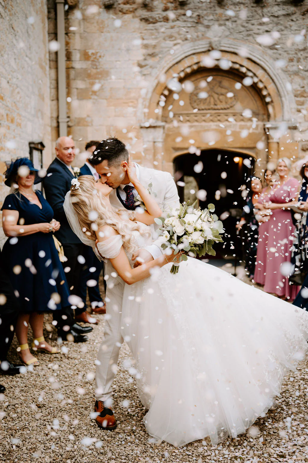 A groom kisses his bride as white confetti swirls in front of a carved stone doorway while guests watch