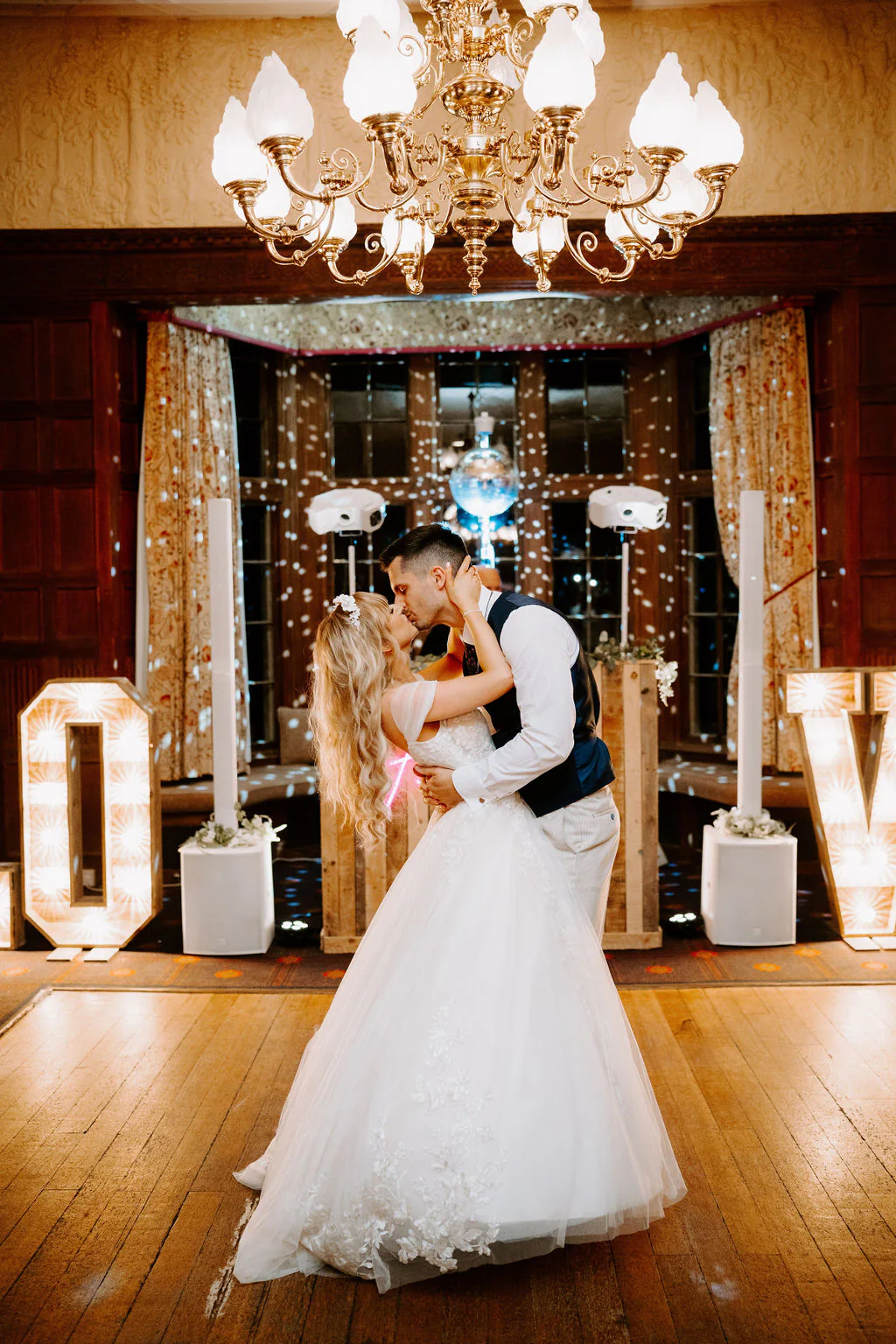 A groom dips his bride and kisses her beneath a chandelier in a dark-panelled room, with big light-up letters and fairy lights behind them