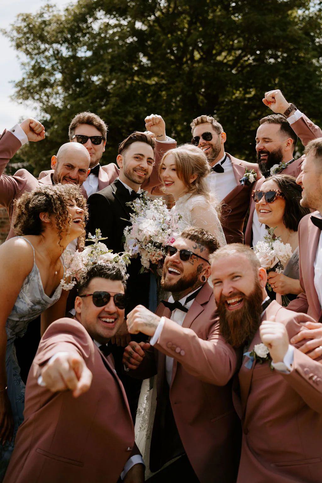 A crowd of friends in dusty pink suits and dresses, many in sunglasses, cheer and point at the camera around a bride holding a bouquet