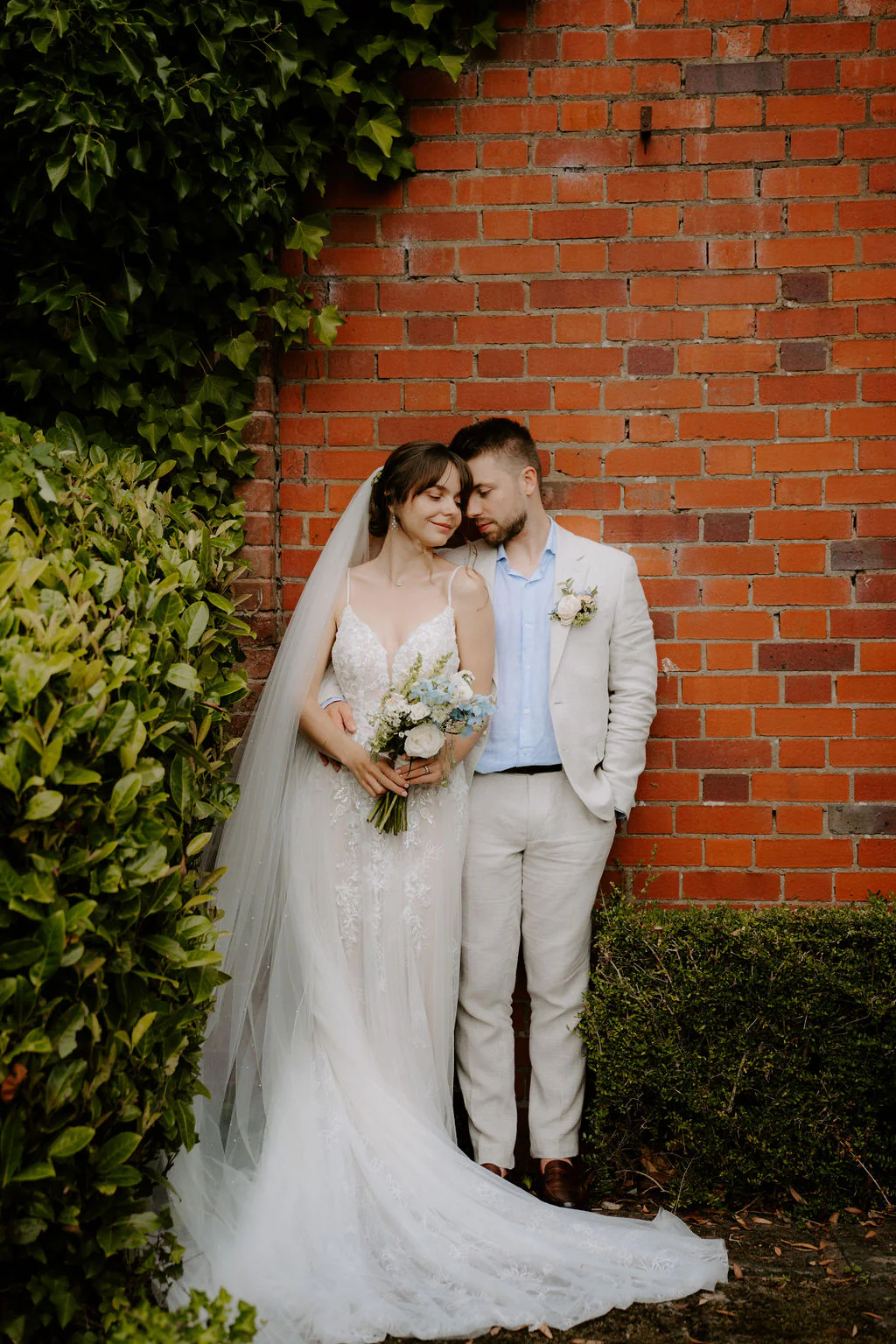 Liz and Jack lean together against a red-brick wall covered in ivy, Liz holding a bouquet of white and pale blue flowers with her veil spread around her feet