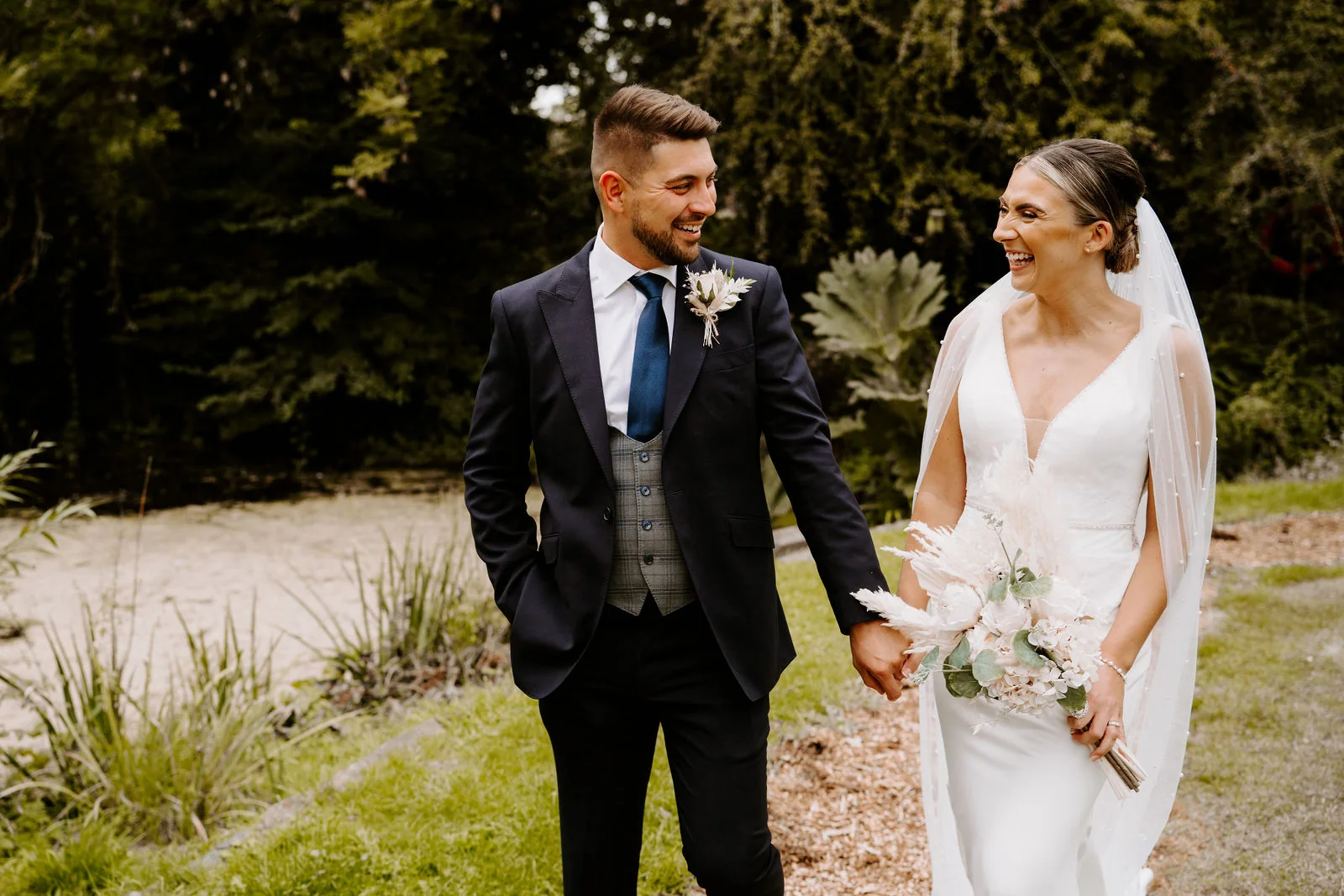 A laughing bride in a veil holding a bouquet of pampas grass and a groom in a navy suit walk hand in hand beside a sandy pond in a garden
