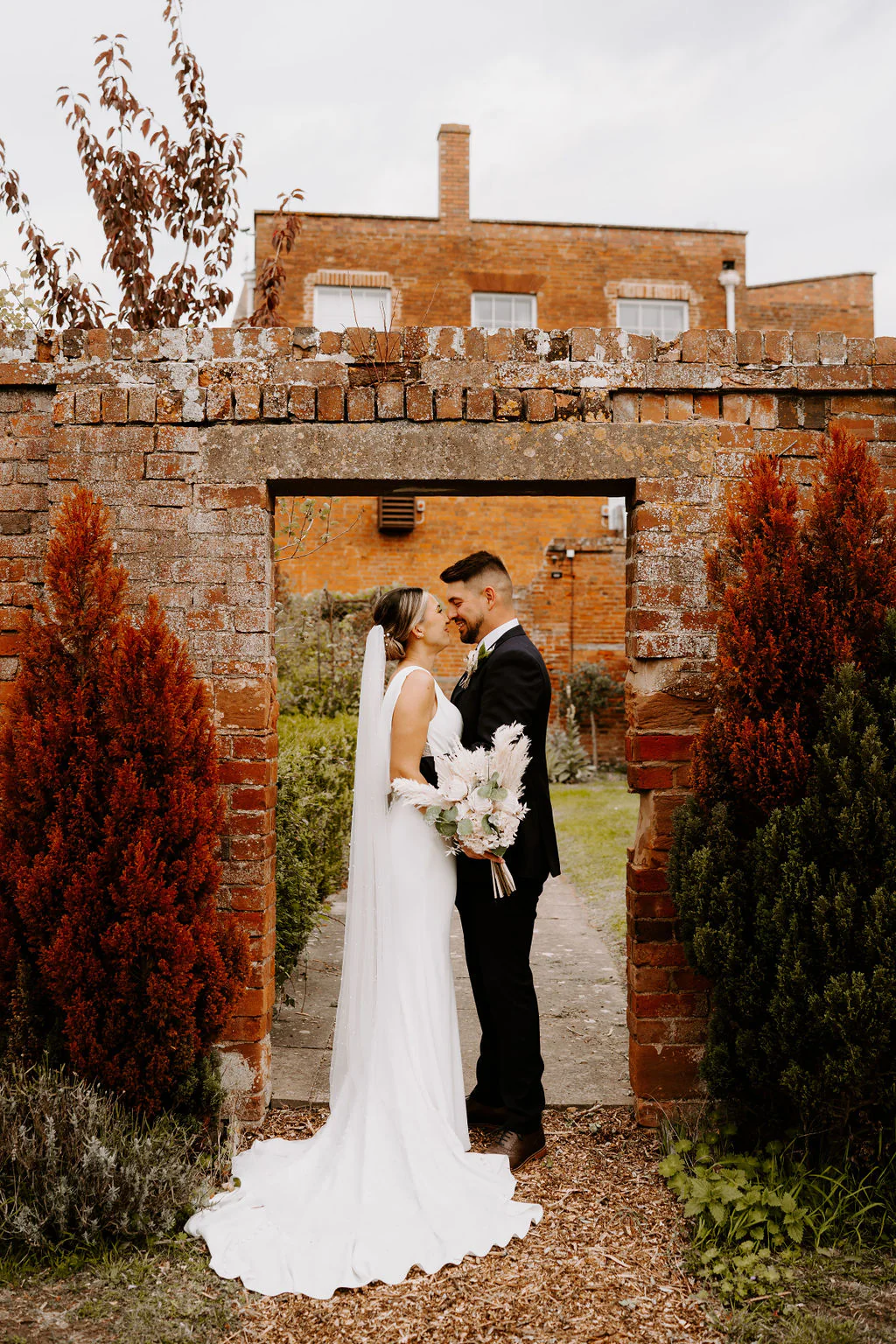 Abi and Adam kiss in a brick archway among red foliage at Stanbrook Abbey Hotel, Abi in a long veil