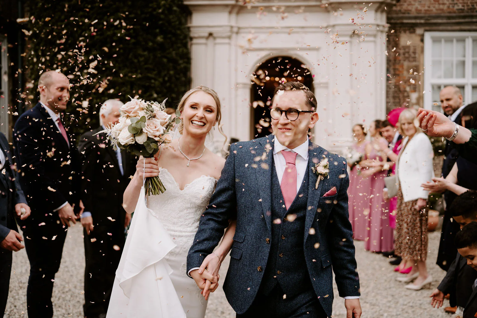 Danielle and Matt, in a navy tweed suit and pink tie, walk hand in hand through confetti thrown by guests in front of the stone porch at Goldsborough Hall