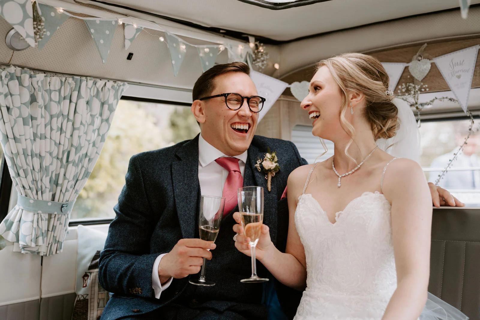 Danielle and Matt laugh and raise glasses of champagne in a room hung with bunting and floral curtains