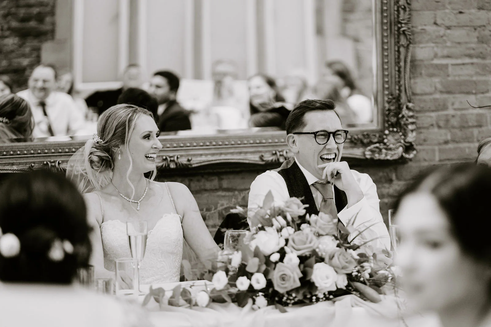 In black and white, Danielle and Matt laugh together at the top table with a bouquet in front of them and a gilt mirror behind