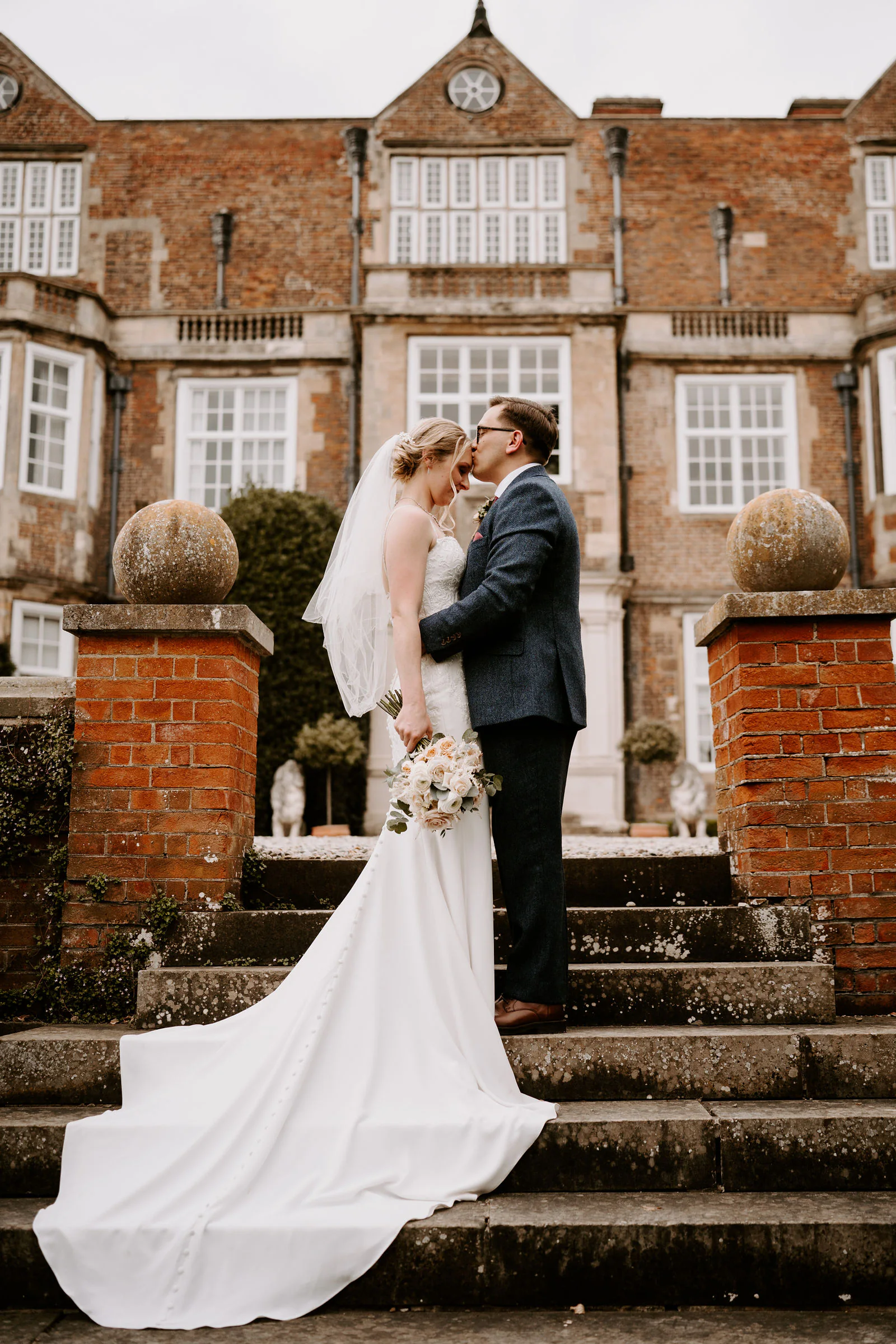 Danielle in a long gown and Matt in a navy suit stand close together on stone steps in front of the red-brick front of Goldsborough Hall, her train spread behind her