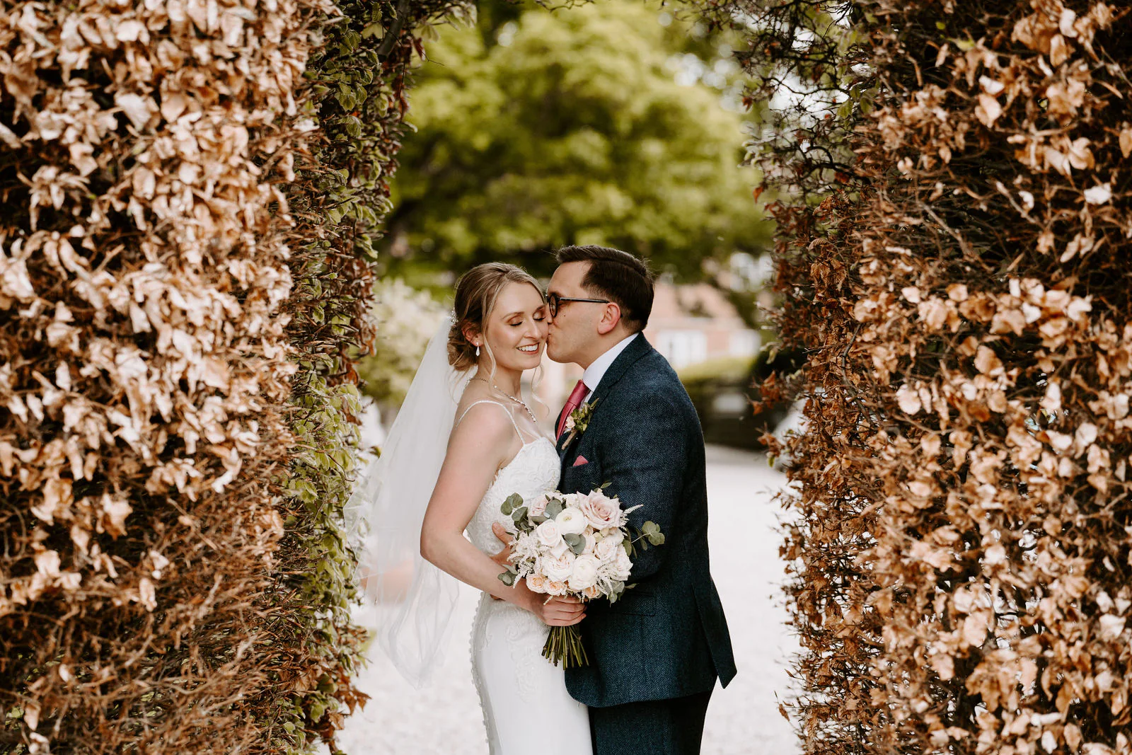 Danielle and Matt kiss cheek to cheek between two tall hedges of russet beech leaves, Danielle holding a bouquet of white flowers