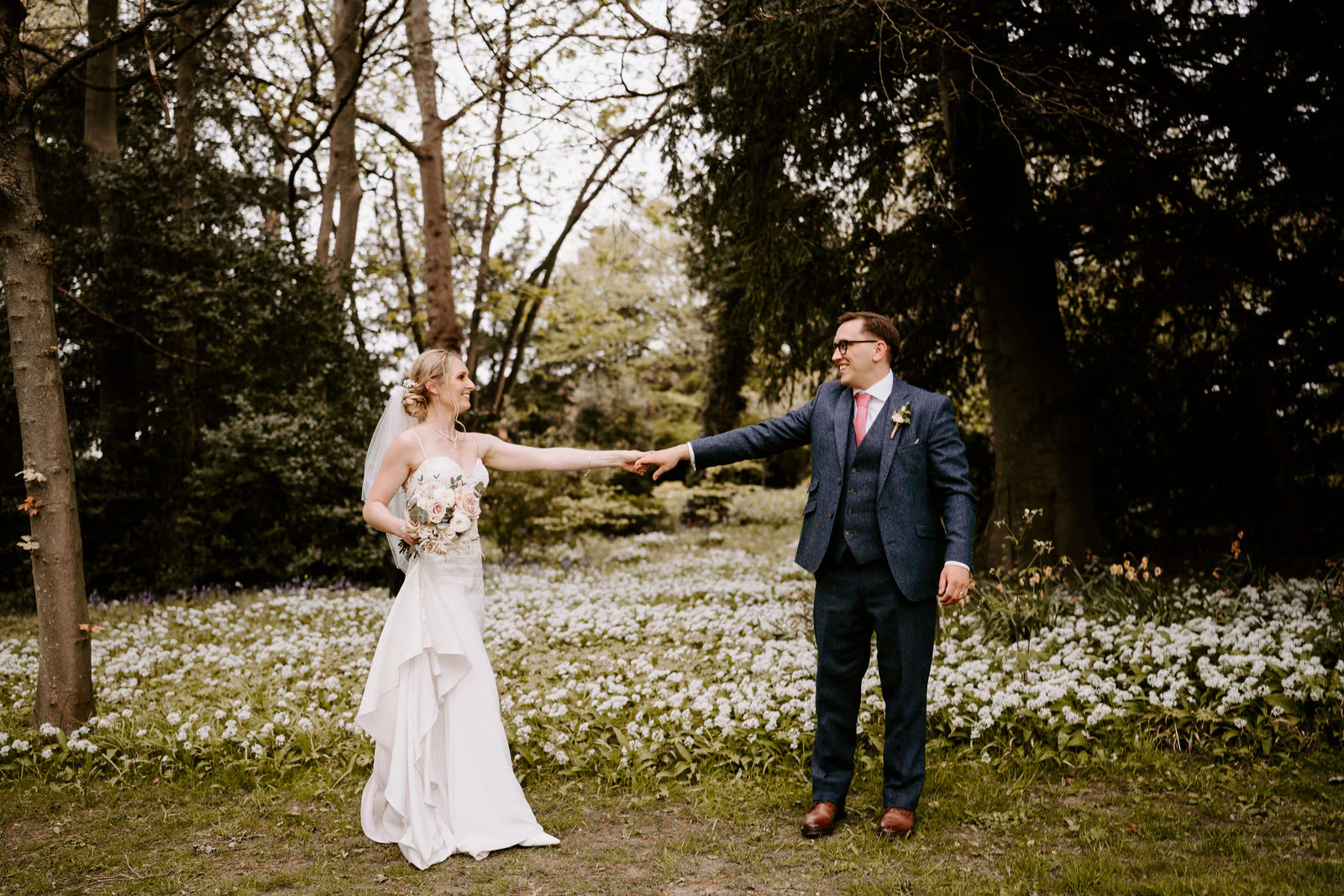 Danielle stretches out her hand to Matt across a woodland lawn carpeted with white flowers, tall trees behind them