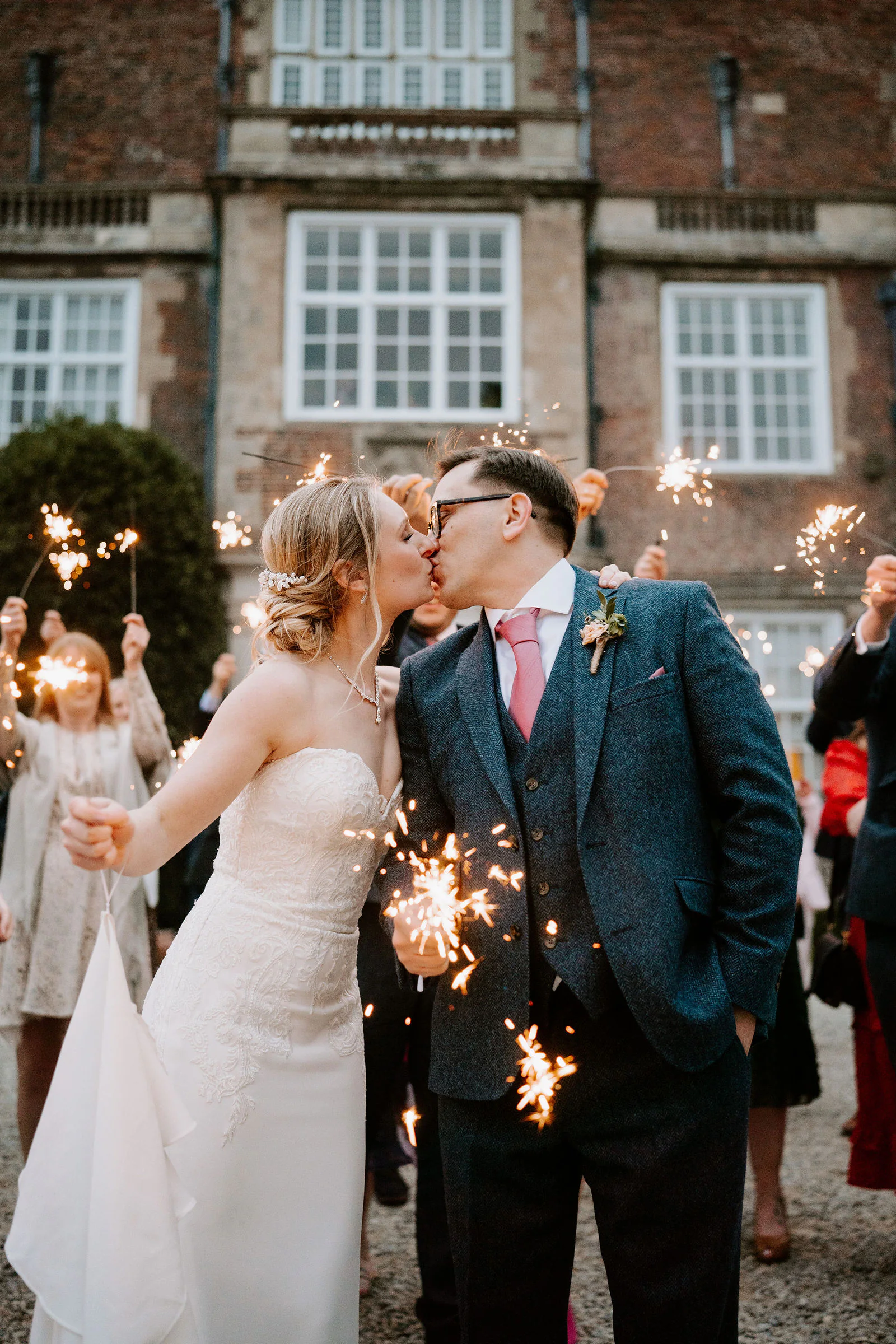 Danielle and Matt kiss as guests wave sparklers around them in front of the stone hall at Goldsborough Hall